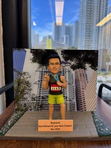 一個人像公仔，身穿馬拉松運動服裝，背號1103，站在模擬比賽場景中，做出勝利手勢。背景是一個城市景觀，展示了摩天大樓和樹木，底座上標有「Raymond Best Wishes for your New Chapter Nov 2024」的祝福語