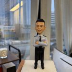 一尊以「督察」職級為主題的警察人像公仔，被手掌托於室內窗邊。公仔短黑髮、微笑神情；身穿淺藍色長袖制服襯衫，配黑色領帶與深色肩章，左胸名牌旁印有「POLICE」字樣；雙臂於胸前交叉。下身為黑色長褲與黑色皮鞋。人物站在淺米色粗糙質感的方形底座上。背景為落地窗外的城市街景：密集高樓、道路與行道樹；玻璃上可見長條燈的反射。室內有米色窗簾、木質小桌與蠟燭杯，以及白色設備邊角。整體呈現在辦公室窗邊展示、便於送禮與陳列的情境。