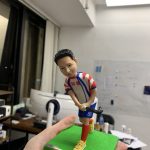 一尊動作姿態的曲棍球人像公仔，站在草綠色方形地台上，雙手握著黑色曲棍球棍，準備擊打腳前的白色球。公仔穿著紅藍白相間的球衣與短褲，胸前與褲上印有數字「80」及圓形隊章樣式，右腿配戴紅色護襪，腳穿黃色球鞋。地台前方名牌寫有「Dr. Alex W.K. CHAN」。背景為室內工作室環境，牆上有白板與貼紙。此作品屬人像公仔與曲棍球人像公仔的訂製運動主題擺設。