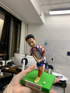 一個名為Dr. Alex W.K. CHAN的曲棍球教練人像公仔，穿著紅白藍相間的曲棍球球衣，背號80，正處於擊球姿勢，腳下是綠色草地底座。