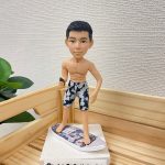 一款滑浪主題的人像公仔站在迷你滑浪板上，呈半轉身站姿。公仔為短黑髮、上身赤裸，穿深藍與灰白花紋的及膝沙灘短褲，赤腳踏在帶有圖案貼膜的白色迷你滑浪板上，手中握著一支小型棕色配件。整體擺設置於淺色木製托盤內，旁邊有綠色植物點綴，背景是米白色細紋牆面；前景可見「Cutefigure_hk」的名牌。此作品為運動主題的滑浪人像公仔與人像公仔。