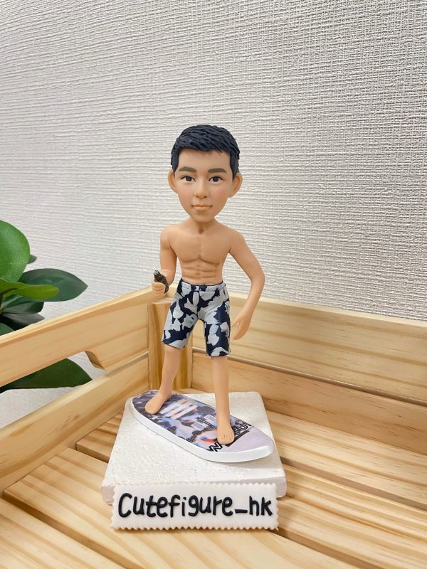 一款滑浪主題的人像公仔站在迷你滑浪板上，呈半轉身站姿。公仔為短黑髮、上身赤裸，穿深藍與灰白花紋的及膝沙灘短褲，赤腳踏在帶有圖案貼膜的白色迷你滑浪板上，手中握著一支小型棕色配件。整體擺設置於淺色木製托盤內，旁邊有綠色植物點綴，背景是米白色細紋牆面；前景可見「Cutefigure_hk」的名牌。此作品為運動主題的滑浪人像公仔與人像公仔。