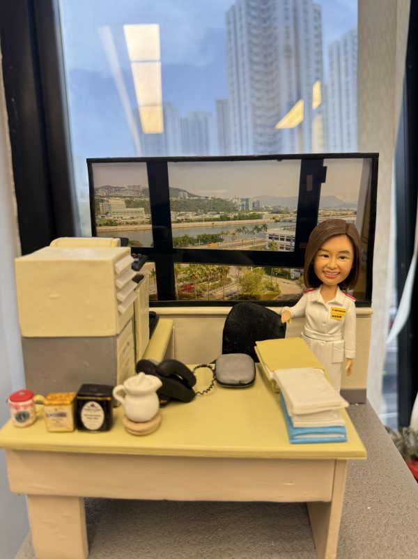 透明展示盒內的辦公室場景人像公仔：一位短髮女性穿白色制服、肩章與名牌，站在米黃色書桌旁微笑；桌上有文件疊、座機電話、滑鼠、茶具與幾罐沖泡品，左側是一部多功能影印／掃描器；背後窗景為臨河城市與山景的大幅相片，呈現醫院或醫療機構行政辦公風格。
