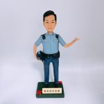 一尊以「警員」職級為主題的人像公仔置於白色無縫背景前拍攝。公仔黑色短髮、微笑神情；身穿淺藍色短袖制服襯衫，肩上有深色肩墊與扣件，左胸口袋上方印有 POLICE 字樣。腰間繫黑色裝備腰帶，配有多個裝備袋與扣具；下身為深藍長褲與黑色皮鞋。右手自然下垂並持一頂深藍色頭盔，左臂抬起向側前方打開手掌做示意/招呼姿態。腳下為深綠色方形台座，前側裝有米金色名牌，刻有「爸爸退休快樂」等祝福字樣，台座兩角點綴紅色小花。整體以簡潔白底呈現，凸顯公仔與制服細節，營造紀念與致意的送禮場景。