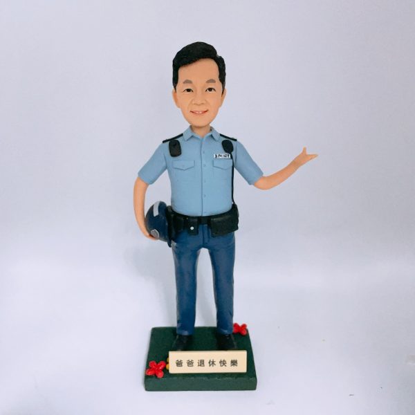 一尊以「警員」職級為主題的人像公仔置於白色無縫背景前拍攝。公仔黑色短髮、微笑神情；身穿淺藍色短袖制服襯衫，肩上有深色肩墊與扣件，左胸口袋上方印有 POLICE 字樣。腰間繫黑色裝備腰帶，配有多個裝備袋與扣具；下身為深藍長褲與黑色皮鞋。右手自然下垂並持一頂深藍色頭盔，左臂抬起向側前方打開手掌做示意/招呼姿態。腳下為深綠色方形台座，前側裝有米金色名牌，刻有「爸爸退休快樂」等祝福字樣，台座兩角點綴紅色小花。整體以簡潔白底呈現，凸顯公仔與制服細節，營造紀念與致意的送禮場景。
