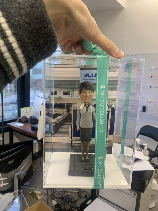 一款為全日空（ANA）航空女地勤人員訂製的人像公仔，公仔身穿制服，被放置在一個透明禮盒中，其背景為機場櫃檯的配件裝飾。