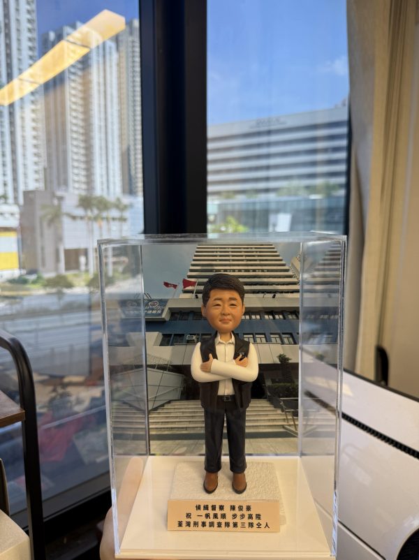 客製化警察人像公仔，身穿白襯衫與黑色背心，雙手交叉微笑站立，底座配有專屬銘牌，背景為警署大樓。銘牌刻有「偵緝警察 陳俊豪 祝一帆風順 步步高陞 荃灣刑事調查隊第三隊全人」字樣，象徵同袍對升職的祝賀與鼓勵。這款警察人像公仔適合用於升職賀禮、榮休紀念或作為警務人員的專屬禮物收藏。