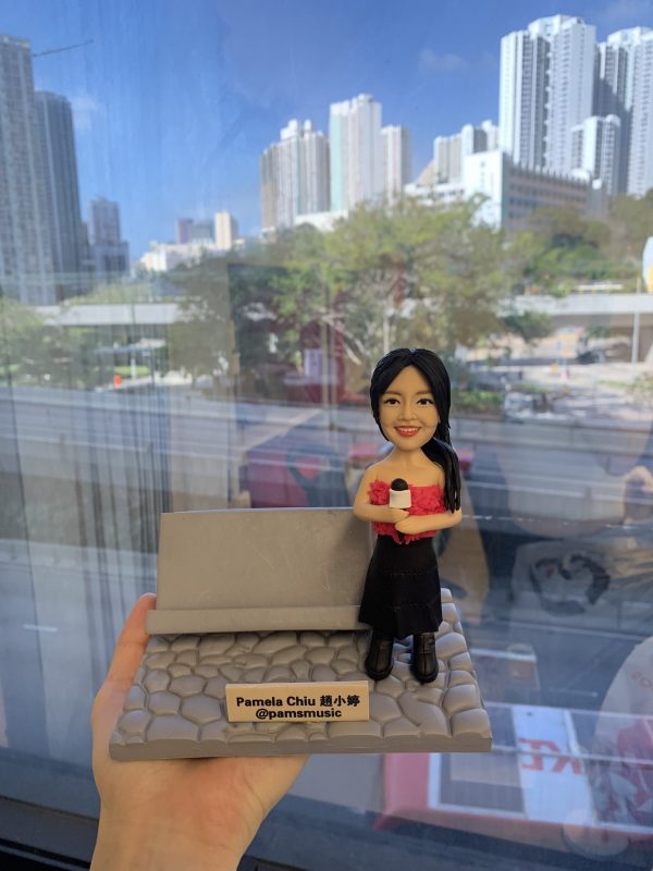 香港歌手趙小婷（Pamela Chiu）的訂製人像公仔，公仔手持咪高峰，站在鵝卵石地台上，其背景配件裝飾為一張灰色長櫈。