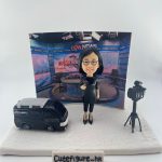 新聞採訪主題人像公仔場景：短髮戴眼鏡的女性記者穿黑色連身裙與高跟鞋，手持麥克風站在白色地台上；左側是一台黑色新聞採訪車模型，右側為架在三腳架上的專業攝影機；背板呈現演播室畫面與節目佈景。整體具專業與儀式感，適合作為新聞記者退休禮物與致敬紀念擺設。