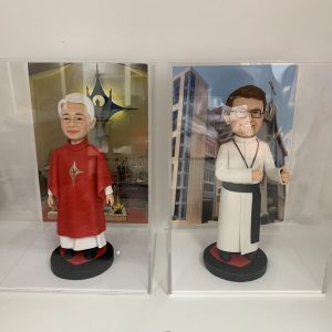 兩款並排展示在透明亞加力膠盒中的客製化神父人像公仔，左邊身穿紅色祭披，右邊身穿白色聖袍比讚，是送給牧師或神職人員的禮物系列。
