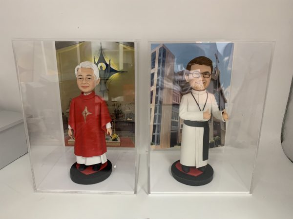 兩款並排展示在透明亞加力膠盒中的客製化神父人像公仔，左邊身穿紅色祭披，右邊身穿白色聖袍比讚，是送給牧師或神職人員的禮物系列。