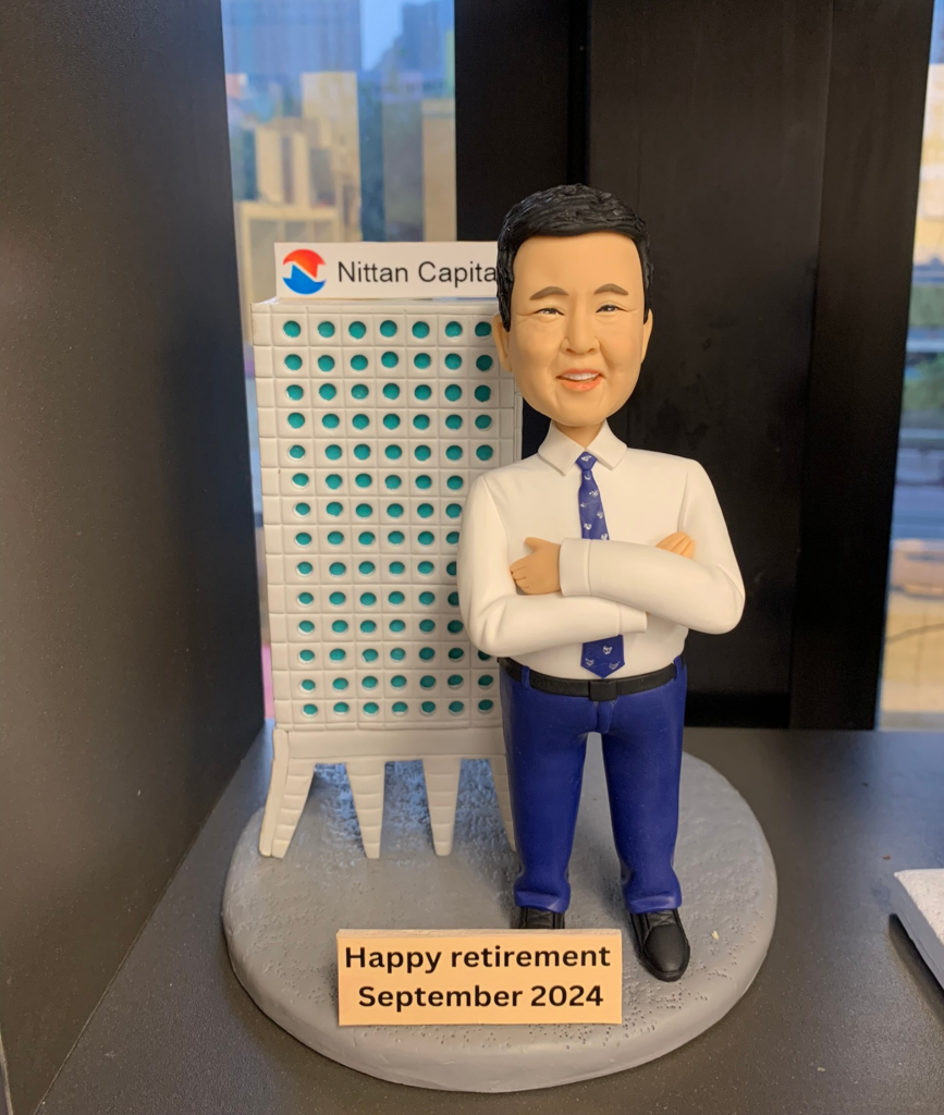 一款為Nittan Capital老闆訂製的退休禮物。精緻的人像公仔雙手抱胸，自信地站在公司大樓模型旁邊，底座銘牌刻有「Happy retirement September 2024」，是一份極具心思的公司禮物，彰顯了對領導者的敬意。
