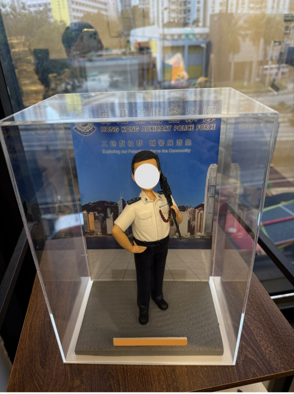 這是一款精心定製的香港警察專屬人像公仔，身穿輔警制服，肩上配有步槍，細節處理極為逼真，從警徽到制服線條均一絲不苟。公仔被安置於透明展示盒中，背景是印有「香港輔助警察隊」字樣與香港天際線的設計，突顯警隊守護社區的責任與使命。整體造型既專業莊嚴，又帶有收藏與紀念的意義，成為一份別具心意的專屬禮物。
