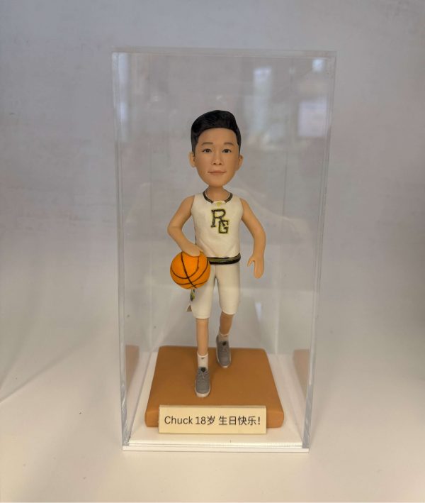 籃球員主題人像公仔：短髮少年公仔身穿白色籃球球衣與球褲，胸前有金邊字母標誌，腳踩灰色球鞋；右手持橙色籃球、左腳邁步向前的運球姿勢。公仔站在橘棕色方形底座上，前方銘牌寫有「Chuck 18岁 生日快乐！」字樣；整體置於透明展示盒中，白色背景簡潔，呈現適合作為生日禮物的籃球主題人像公仔。