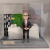 CEO主題人像公仔退休禮物擺設：灰白短髮、戴眼鏡的男士公仔身穿深色西裝與條紋領帶，雙手於腹前自然交握微笑站立。場景為公司前台辦公區造型：幾何白色接待櫃台、右側背板印有企業Logo字樣，左後方有黑色書櫃與書本、中央為綠色植栽牆，櫃台上擺放白色花盆植栽與小方形品牌色塊。地面為灰色地毯質感底板，前側附金屬色名牌刻有任期資訊。作品可放入透明展示盒收藏，整體呈現向老闆與CEO致意的退休禮物人像公仔。