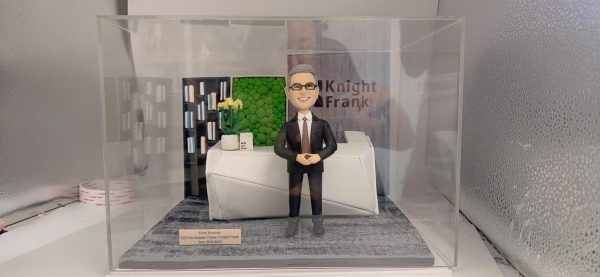 CEO主題人像公仔退休禮物擺設：灰白短髮、戴眼鏡的男士公仔身穿深色西裝與條紋領帶，雙手於腹前自然交握微笑站立。場景為公司前台辦公區造型：幾何白色接待櫃台、右側背板印有企業Logo字樣，左後方有黑色書櫃與書本、中央為綠色植栽牆，櫃台上擺放白色花盆植栽與小方形品牌色塊。地面為灰色地毯質感底板，前側附金屬色名牌刻有任期資訊。作品可放入透明展示盒收藏，整體呈現向老闆與CEO致意的退休禮物人像公仔。