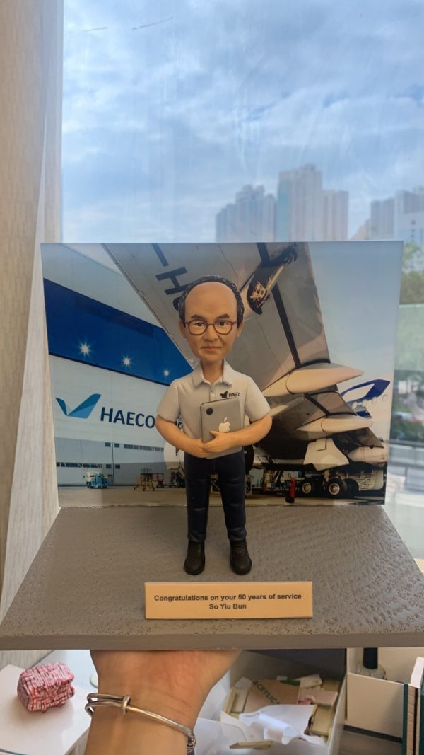 人像公仔退休禮物：中年男士公仔留短髮、戴黑框眼鏡，穿淺灰色短袖襯衫（左胸口繡有 HAECO 標誌）與深色長褲、黑色工作靴，雙手抱著貼有蘋果貼紙的平板電腦，站在灰色質感底座上；前方金色名牌印有「Congratulations on your 50 years of service / So Yiu Bun」。背板為停機坪與大型客機機翼、機庫外牆與 HAECO 字樣的場景，展現專業維修或營運背景。作品於室內靠窗拍攝，畫面下方可見手掌托住底座。