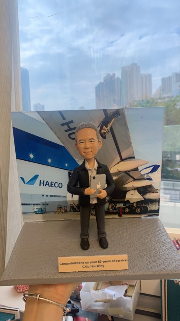 人像公仔退休禮物：中年男士公仔留短髮、戴黑框眼鏡，穿淺灰色短袖襯衫（左胸口繡有 HAECO 標誌）與深色長褲、黑色工作靴，雙手抱著貼有蘋果貼紙的平板電腦，站在灰色質感底座上；前方金色名牌印有「Congratulations on your 50 years of service / So Yiu Bun」。背板為停機坪與大型客機機翼、機庫外牆與 HAECO 字樣的場景，展現專業維修或營運背景。作品於室內靠窗拍攝，畫面下方可見手掌托住底座。
