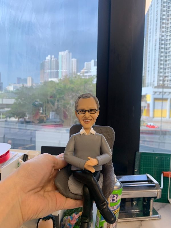 CEO主題人像公仔：灰白短髮、戴眼鏡的男士公仔坐在深灰色紋理單人椅上，雙手自然交疊放於膝上、雙腿交叉，穿淺灰色針織上衣與黑色長褲、黑色皮鞋，面帶微笑。畫面於室內靠窗拍攝，窗外可見高樓大廈與街景；前景有手掌托住座椅，桌面上可見文具與工具罐，呈現寫實辦公氛圍，適合作為老闆或CEO的退休禮物人像公仔。