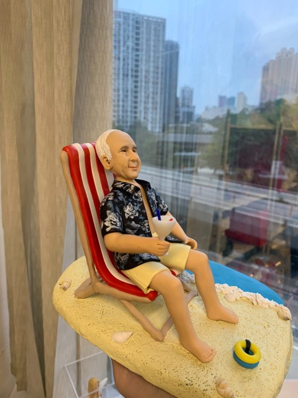 沙灘主題的人像公仔退休禮物：中年至長者男性公仔留短髮、面帶微笑，穿著黑底白花的夏威夷襯衫與米黃色短褲，赤腳坐在紅白條紋海灘椅上，右手握著帶藍色吸管與檸檬片的雞尾酒杯；腳邊細緻呈現米色沙灘、白色浪花邊與貝殼裝飾，旁有黃藍色救生圈小物。底座呈長形沙洲延伸至藍色海面，整體氛圍開心放鬆。拍攝於室內靠窗，背景可見城市高樓與道路景色，近距離與側拍角度皆強調度假感。