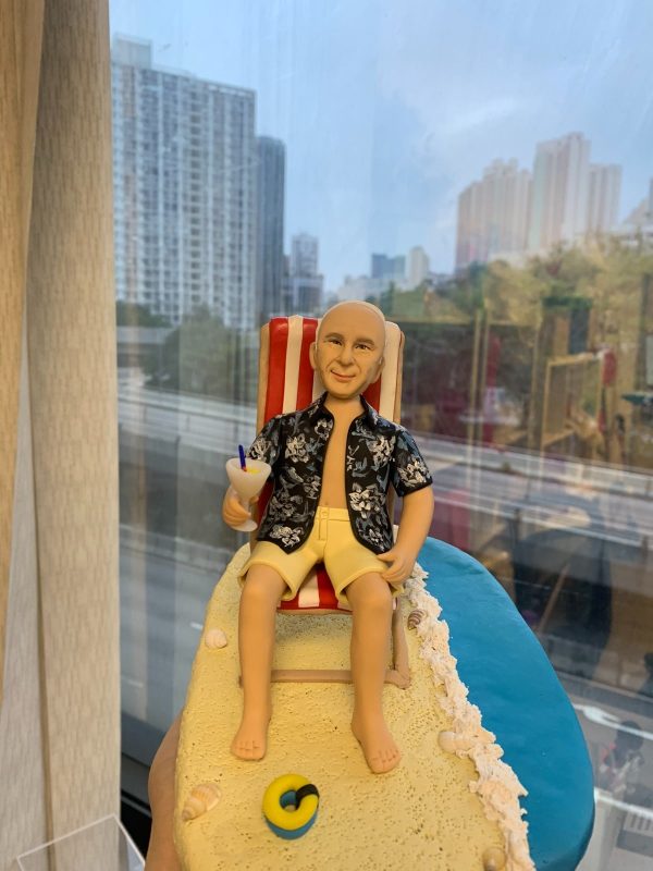 沙灘主題的人像公仔退休禮物：中年至長者男性公仔留短髮、面帶微笑，穿著黑底白花的夏威夷襯衫與米黃色短褲，赤腳坐在紅白條紋海灘椅上，右手握著帶藍色吸管與檸檬片的雞尾酒杯；腳邊細緻呈現米色沙灘、白色浪花邊與貝殼裝飾，旁有黃藍色救生圈小物。底座呈長形沙洲延伸至藍色海面，整體氛圍開心放鬆。拍攝於室內靠窗，背景可見城市高樓與道路景色，近距離與側拍角度皆強調度假感。