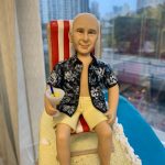 沙灘主題的人像公仔退休禮物：中年至長者男性公仔留短髮、面帶微笑，穿著黑底白花的夏威夷襯衫與米黃色短褲，赤腳坐在紅白條紋海灘椅上，右手握著帶藍色吸管與檸檬片的雞尾酒杯；腳邊細緻呈現米色沙灘、白色浪花邊與貝殼裝飾，旁有黃藍色救生圈小物。底座呈長形沙洲延伸至藍色海面，整體氛圍開心放鬆。拍攝於室內靠窗，背景可見城市高樓與道路景色，近距離與側拍角度皆強調度假感。