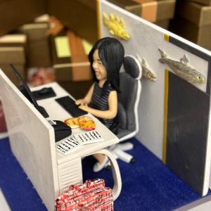 辦公室人像公仔榮休主題:黑長髮女士公仔坐在黑白色辦公椅上,面帶微笑於白色L形辦公桌前工作;桌上有黑色螢幕、鍵盤、滑鼠,旁放開啟的文件與便條,以及兩包金黃色點心。桌側地面放著紅白格紋旅行袋;工位隔板為白灰配色並貼有立體魚造型裝飾(含金色與銀色魚),後方牆面有黑邊與黃條設計。地台為深藍色質感,背景可見成堆咖啡色禮盒與緞帶,營造歡送與祝賀的氛圍。整體呈現細節豐富、帶有個人化元素的榮休辦公室人像公仔,適合作為退休禮物。
