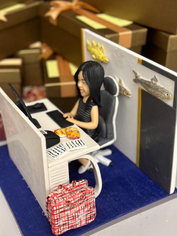 辦公室人像公仔榮休主題：黑長髮女士公仔坐在黑白色辦公椅上，面帶微笑於白色L形辦公桌前工作；桌上有黑色螢幕、鍵盤、滑鼠，旁放開啟的文件與便條，以及兩包金黃色點心。桌側地面放著紅白格紋旅行袋；工位隔板為白灰配色並貼有立體魚造型裝飾（含金色與銀色魚），後方牆面有黑邊與黃條設計。地台為深藍色質感，背景可見成堆咖啡色禮盒與緞帶，營造歡送與祝賀的氛圍。整體呈現細節豐富、帶有個人化元素的榮休辦公室人像公仔，適合作為退休禮物。