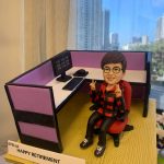 辦公室人像公仔榮休禮：短髮、配圓框眼鏡的上層員工公仔坐在紅色辦公椅上，微笑並以雙手比出雙V手勢；穿紅黑格紋連帽外套、黑色長褲與運動鞋。個人工位為L形配置，粉紫色隔板、白色桌面，配有雙螢幕（畫面顯示「FTE」字樣）、鍵盤、滑鼠與黑色桌上型電話；地台為淺木紋質感。前方白色名牌印有「STELLA｜HAPPY RETIREMENT｜YOUR GS COLLEAGUES｜2023」。作品於室內靠窗拍攝，窗外是晴朗藍天與城市高樓，整體呈現活力、親切且富儀式感的榮休主題辦公室人像公仔。