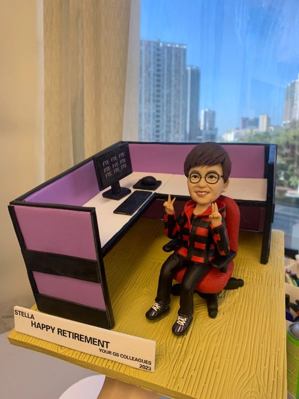 辦公室人像公仔榮休禮：短髮、配圓框眼鏡的上層員工公仔坐在紅色辦公椅上，微笑並以雙手比出雙V手勢；穿紅黑格紋連帽外套、黑色長褲與運動鞋。個人工位為L形配置，粉紫色隔板、白色桌面，配有雙螢幕（畫面顯示「FTE」字樣）、鍵盤、滑鼠與黑色桌上型電話；地台為淺木紋質感。前方白色名牌印有「STELLA｜HAPPY RETIREMENT｜YOUR GS COLLEAGUES｜2023」。作品於室內靠窗拍攝，窗外是晴朗藍天與城市高樓，整體呈現活力、親切且富儀式感的榮休主題辦公室人像公仔。