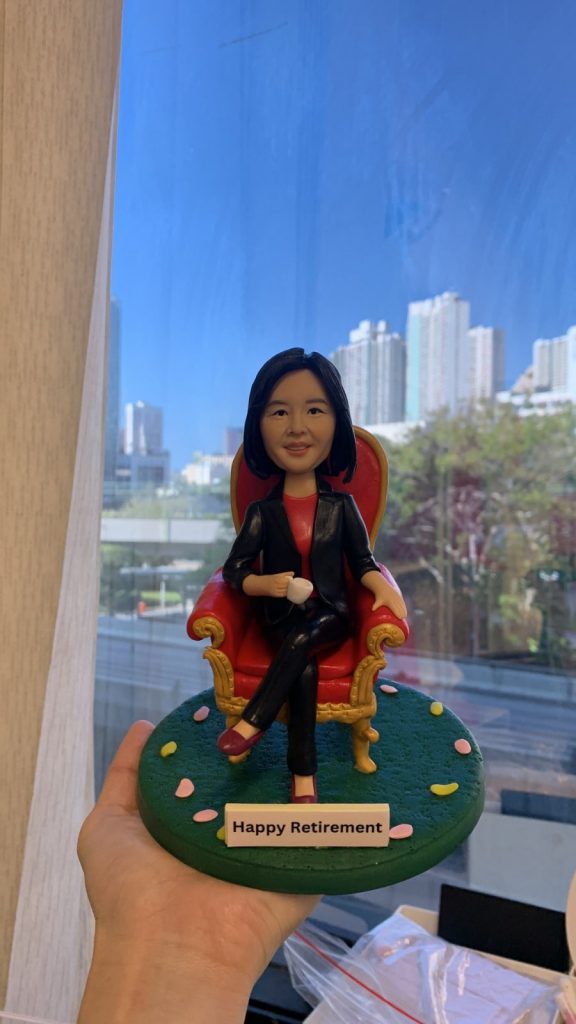 辦公室主題的人像公仔退休禮物：短髮女老闆公仔面帶微笑，穿黑白花紋無袖上衣，坐在黑色辦公椅後、深棕色木質辦公桌前。桌面配置黑色鍵盤、滑鼠、雙螢幕與桌燈，桌側有白色複合式影印機/印表機細節。背景為辦公室玻璃隔間與百葉窗，地台為灰色質感地面，前方小名牌印有「Morgan Stanley」。整體封裝成場景式模型，於室內靠窗拍攝，窗外可見高樓城市景。作品呈現專業而溫暖的榮退氛圍，適合作為老闆的退休禮物人像公仔。