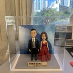雙人款人像公仔退休禮物：一位男士公仔穿黑色西裝、白襯衫與黑領帶，搭配黑皮鞋；旁邊女士公仔穿深紅色V領層次晚禮服，配戴珍珠項鍊，髮型為齊肩短髮。兩人並肩站立於灰色質感底座上，前方金色名牌刻有「EA GROUP CELLQUEEN」與中文公司名稱。背板印製現代商業建築與公司字樣，整體封裝於透明壓克力展示盒中；拍攝於室內靠窗，窗外可見高樓與晴朗藍天。作品呈現正式而隆重的慶賀氛圍，適合作為致敬老闆的退休禮物人像公仔。