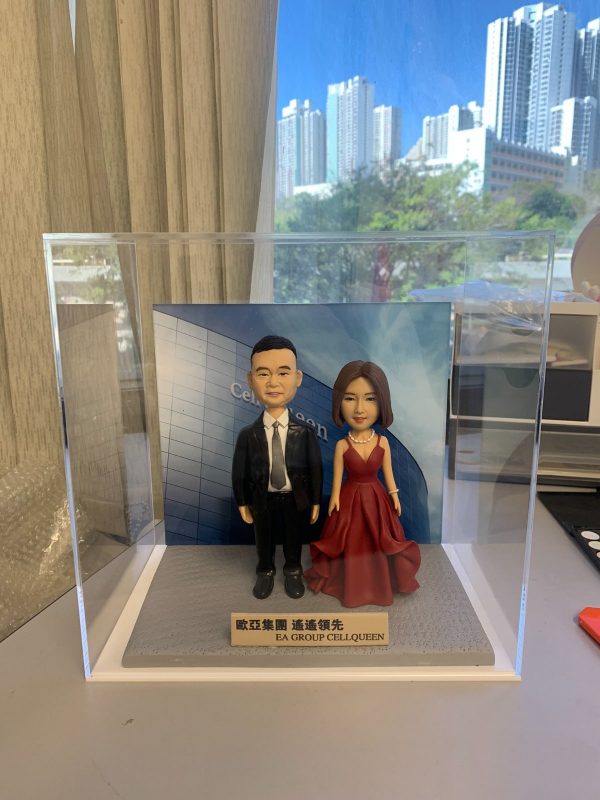 雙人款人像公仔退休禮物：一位男士公仔穿黑色西裝、白襯衫與黑領帶，搭配黑皮鞋；旁邊女士公仔穿深紅色V領層次晚禮服，配戴珍珠項鍊，髮型為齊肩短髮。兩人並肩站立於灰色質感底座上，前方金色名牌刻有「EA GROUP CELLQUEEN」與中文公司名稱。背板印製現代商業建築與公司字樣，整體封裝於透明壓克力展示盒中；拍攝於室內靠窗，窗外可見高樓與晴朗藍天。作品呈現正式而隆重的慶賀氛圍，適合作為致敬老闆的退休禮物人像公仔。