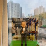 倉庫主題的人像公仔退休禮物：灰白短髮男士公仔表情沉穩、雙手抱胸，身穿深灰色西裝與白襯衫、黑領帶，站在草綠色質感底座上；前方名牌印有「Sing Gor｜Happy Retirement 2024｜身體健康 享樂人生」。背板為高層貨架與堆疊紙箱的現代倉庫場景，可見堆高機與棧板細節；底座左下擺放小石子，右下有編織籃裝綠色果實的裝飾物。作品於室內靠窗拍攝，窗外是城市高樓景色，整體呈現專業與祝賀並重的老闆退休紀念人像公仔。