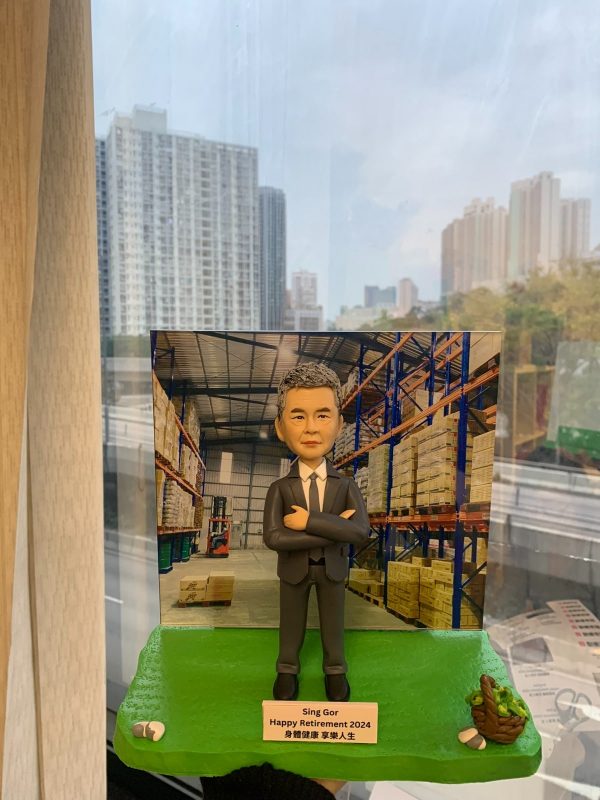 倉庫主題的人像公仔退休禮物：灰白短髮男士公仔表情沉穩、雙手抱胸，身穿深灰色西裝與白襯衫、黑領帶，站在草綠色質感底座上；前方名牌印有「Sing Gor｜Happy Retirement 2024｜身體健康 享樂人生」。背板為高層貨架與堆疊紙箱的現代倉庫場景，可見堆高機與棧板細節；底座左下擺放小石子，右下有編織籃裝綠色果實的裝飾物。作品於室內靠窗拍攝，窗外是城市高樓景色，整體呈現專業與祝賀並重的老闆退休紀念人像公仔。