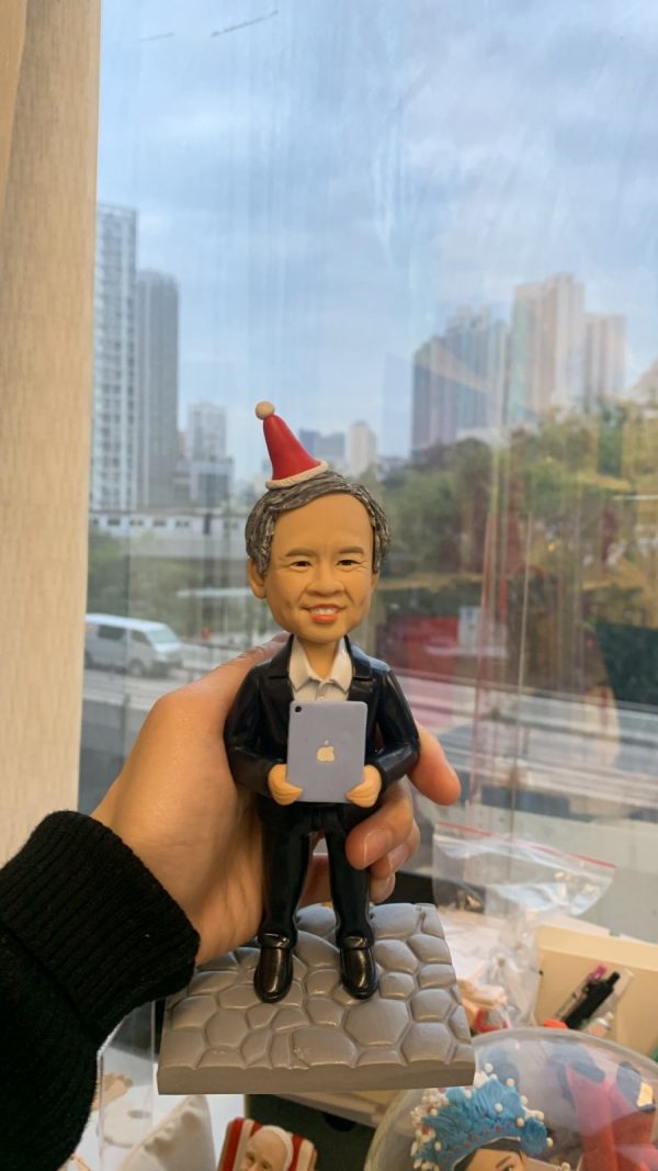 人像公仔退休禮物：中年至長者男士公仔留短灰髮、面帶微笑，頭戴紅白相間的派對尖帽；身穿深色西裝外套與白襯衫，雙手抱著帶有蘋果圖案的小平板電腦。公仔站在灰色石板紋底座上，由手掌托起於室內靠窗拍攝，窗外可見城市高樓與道路景致；畫面下方有辦公室文具與展示物，呈現輕鬆又正式的慶祝氛圍，適合作為老闆的退休禮物人像公仔。