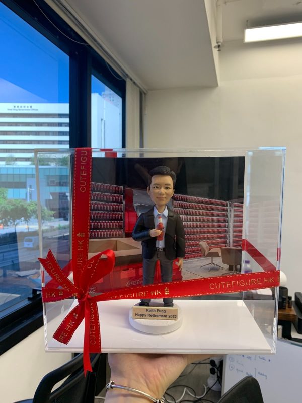 企業主題的人像公仔退休禮物：短髮男士公仔身穿深色西裝外套、白襯衫與紅色領帶，右手拿著一瓶紅色標誌的小汽水；公仔站在圓形白色底座上，底座前方名牌刻有「Keith Fung｜Happy Retirement 2023」。背景板為紅白相間的品牌展示牆與辦公室休憩區場景，前景以透明壓克力展示盒包裹，外繫紅色緞帶與金字「CUTEFIGURE HK」綁帶蝴蝶結；作品由手掌托起拍攝，畫面左側可見大面窗景與城市建築。