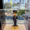 透明壓克力展示盒內擺放一尊以「高級督察」職級為主題的警察人像公仔。公仔頭戴黑色禮帽，帽徽與銀色帽帶清晰；短髮、戴黑框眼鏡，微笑神情。上身著黑色禮服外套，胸前有勳略與綬帶金屬件，肩章與金屬鈕釦立體分明；腰間繫藍色禮儀腰帶，垂掛藍色流蘇。右手自然下垂戴白手套，左手以白手套握於腹前，展現正式儀典姿態；下身為整齊的黑色禮服長褲與黑色禮鞋。公仔立於圓形深灰台座，前方擺放金色名牌，上有致意與日期文字。展示盒內背板是一幅在戶外操場／禮台前的隊列合照，多名著深藍禮服的同袍整齊就座，後方可見校舍或訓練設施與旗桿。展示盒外的環境位於窗邊，窗外是高密度住宅大廈與藍天，室內長條燈具倒影映在玻璃上；整體呈現典禮後的紀念展示與贈禮情境。