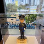 透明壓克力展示盒內擺放一尊以「高級督察」職級為主題的警察人像公仔。公仔頭戴黑色禮帽，帽徽與銀色帽帶清晰；短髮、戴黑框眼鏡，微笑神情。上身著黑色禮服外套，胸前有勳略與綬帶金屬件，肩章與金屬鈕釦立體分明；腰間繫藍色禮儀腰帶，垂掛藍色流蘇。右手自然下垂戴白手套，左手以白手套握於腹前，展現正式儀典姿態；下身為整齊的黑色禮服長褲與黑色禮鞋。公仔立於圓形深灰台座，前方擺放金色名牌，上有致意與日期文字。展示盒內背板是一幅在戶外操場／禮台前的隊列合照，多名著深藍禮服的同袍整齊就座，後方可見校舍或訓練設施與旗桿。展示盒外的環境位於窗邊，窗外是高密度住宅大廈與藍天，室內長條燈具倒影映在玻璃上；整體呈現典禮後的紀念展示與贈禮情境。