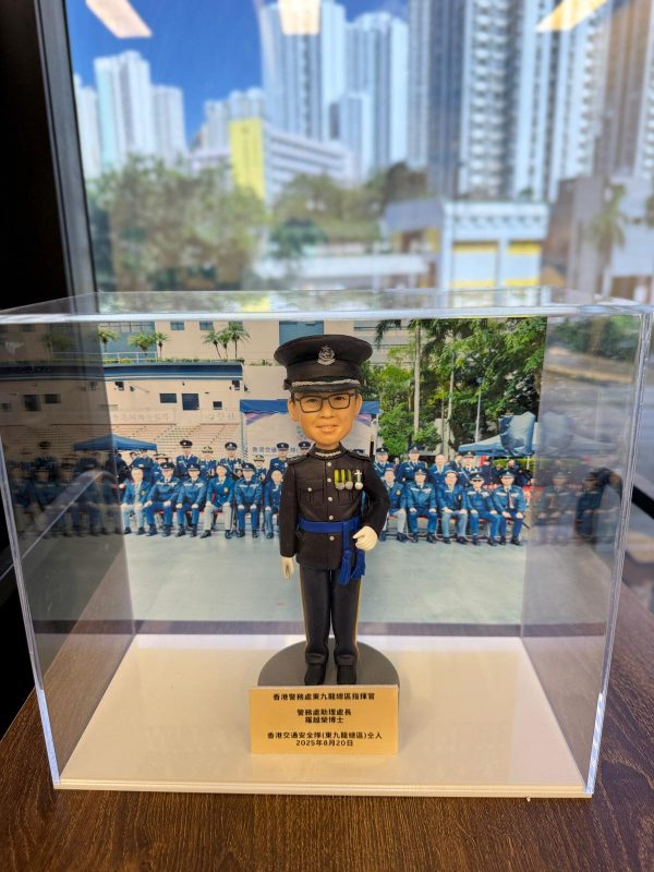 透明壓克力展示盒內擺放一尊以「高級督察」職級為主題的警察人像公仔。公仔頭戴黑色禮帽，帽徽與銀色帽帶清晰；短髮、戴黑框眼鏡，微笑神情。上身著黑色禮服外套，胸前有勳略與綬帶金屬件，肩章與金屬鈕釦立體分明；腰間繫藍色禮儀腰帶，垂掛藍色流蘇。右手自然下垂戴白手套，左手以白手套握於腹前，展現正式儀典姿態；下身為整齊的黑色禮服長褲與黑色禮鞋。公仔立於圓形深灰台座，前方擺放金色名牌，上有致意與日期文字。展示盒內背板是一幅在戶外操場／禮台前的隊列合照，多名著深藍禮服的同袍整齊就座，後方可見校舍或訓練設施與旗桿。展示盒外的環境位於窗邊，窗外是高密度住宅大廈與藍天，室內長條燈具倒影映在玻璃上；整體呈現典禮後的紀念展示與贈禮情境。