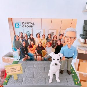 一座客製化場景人像公仔：灰髮眼鏡男子身著淺藍色襯衫與卡其長褲，站在灰色磚面平台上，身旁有一隻白色擬真狗狗。平台右側以草地與花瓣點綴，左前方立有小木牌，上印英文祝福字樣。後方豎立一幅團隊合影相片背景，左上可見「CAPITAL GROUP」字樣與幾何圖形標記。整體呈現溫馨且專業的退休紀念主題，適合作為全球性金融服务公司員工的致敬禮物與商務場合展示的人像公仔作品。