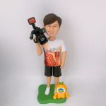 電視台攝錄師主題人像公仔：短髮男子面帶笑容，穿白底橙紅圖案T恤與黑色短褲、白色球鞋，右肩扛專業攝影機，站在綠色草地造型底座上；腳邊有一隻黃色吉祥物小公仔，胸前圓形多色標誌醒目。整體呈現輕鬆又專業的攝錄師形象，適合作為電視台攝錄師退休紀念。