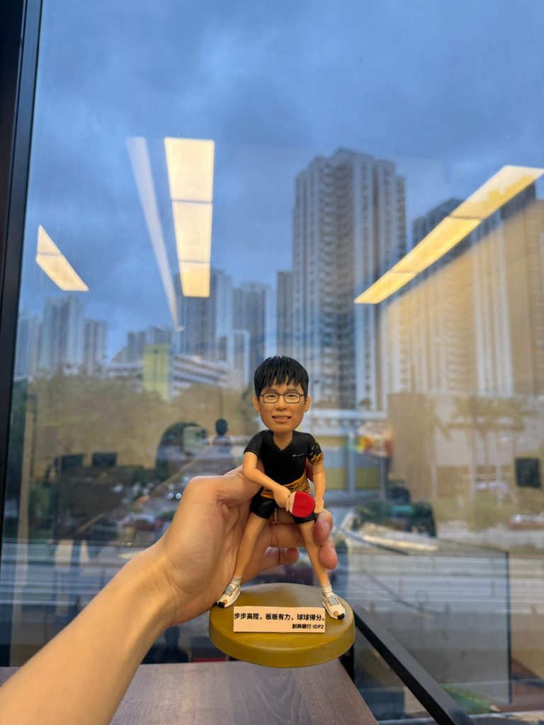 乒乓球比賽主題人像公仔：短髮戴眼鏡的男子公仔身穿黑色運動上衣與短褲，袖口與腰間有金色圖樣點綴；他採低重心準備姿勢，右手握紅色乒乓球拍、左手持白色乒乓球，腳穿白色運動鞋，站在半透明金黃色圓形底座上，底座前貼有中文勵志銘牌。畫面由室內對著落地窗拍攝，可見城市高樓與室內燈光倒影，手持者的手從左側托住公仔。