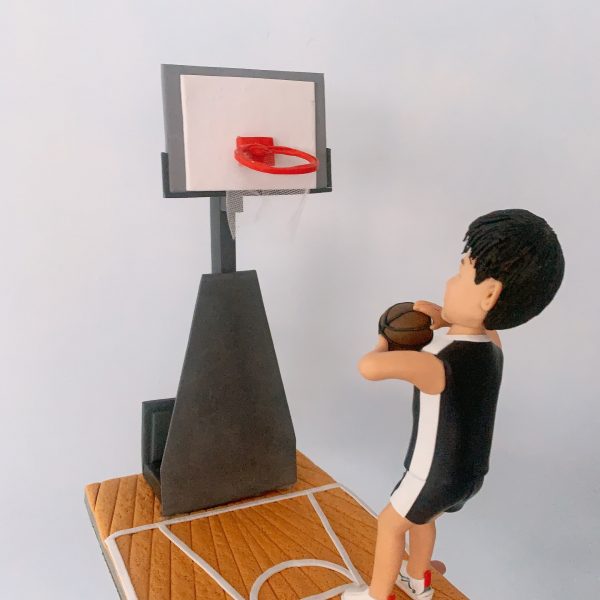 香港訂做籃球運動員公仔，人物着黑白色籃球服短褲，腳踏白色運動鞋，雙手抱住棕色籃球，站在木紋球場底座上，面向紅色籃球架準備投籃。