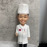 廚師主題人像公仔特寫：戴白色高帽、穿白色廚師袍的男廚師微笑站立，左胸有紅色小旗與三顆星繡章，右胸口有吉祥物圖案；大衣下擺印有「Matsu San」字樣。公仔站在灰色石磚紋底座上，前方白色名牌印「Shikon Yau Yue」。背景為灰色水泥紋牆面與白色紗網質感襯底，前景有綠色底板與黃色、粉色小裝飾。整體呈現隆重且具紀念感，適合作為廚師退休禮物。