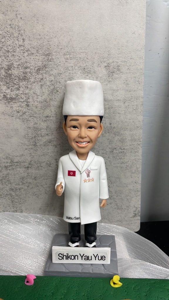 廚師主題人像公仔特寫：戴白色高帽、穿白色廚師袍的男廚師微笑站立，左胸有紅色小旗與三顆星繡章，右胸口有吉祥物圖案；大衣下擺印有「Matsu San」字樣。公仔站在灰色石磚紋底座上，前方白色名牌印「Shikon Yau Yue」。背景為灰色水泥紋牆面與白色紗網質感襯底，前景有綠色底板與黃色、粉色小裝飾。整體呈現隆重且具紀念感，適合作為廚師退休禮物。