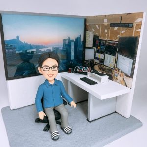 一座辦公室情境的人像公仔：短髮戴眼鏡的男子穿藍色襯衫與灰色長褲，腳踩黑白條紋拖鞋，坐在黑色旋轉椅上微笑。右側是一張白色轉角辦公桌，桌面放有遙控器、長條鍵盤/控制器與支架；背牆貼有交易室環境與多屏幕行情介面照片，左側背景則是城市與海灣天際線的照片。整體置於灰色地台，呈現現代辦公室與金融交易工作氛圍的人像公仔擺設。