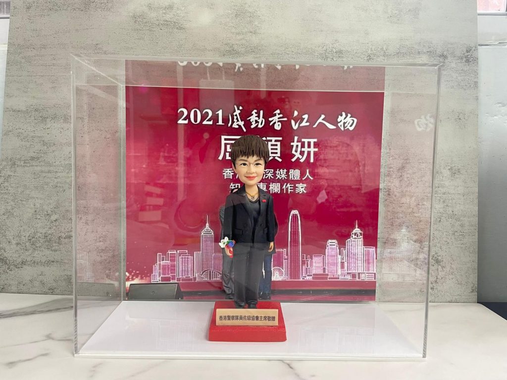 透明亞加力展示盒內的一款精緻手工人像公仔，造型為短髮男子穿深色西裝配黑皮鞋與白恤衫，右手持小花束，站在紅色台座上；台座前方有金色名牌，顯示為香港警隊致贈的禮品（為保護個人識別，名牌人名不展示）。背板為紅色舞台／頒獎設計，上有「2021 感動香江人物」等白色字樣與「資深媒體人、作家」字句，下方繪有香港天際線線條圖。場景置於石紋枱面與灰色牆面前。公仔雕工細膩、五官傳神、塗裝乾淨，整體呈現隆重致敬氛圍。
