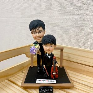 一款音樂老師與學生的雙人訂製人像公仔。小男孩身穿禮服手持大提琴，男老師站在身後拿著花束。公仔地台上有一塊寫上名字的銘牌。