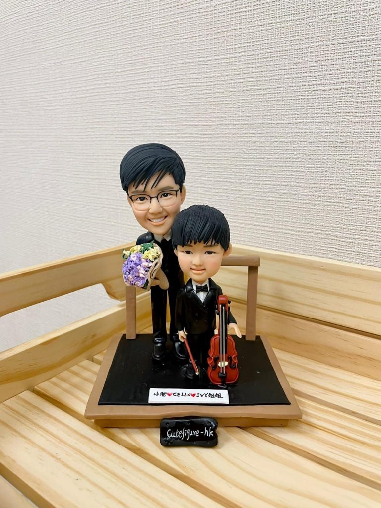 一款音樂老師與學生的雙人訂製人像公仔。小男孩身穿禮服手持大提琴，男老師站在身後拿著花束。公仔地台上有一塊寫上名字的銘牌。